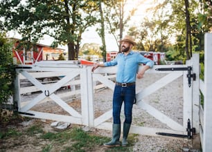 Um retrato do agricultor em pé ao ar livre na fazenda da família, de pé ao lado do portão de madeira.