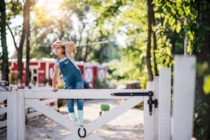 A portrait of happy small girl on family farm, standing by wooden gate.
