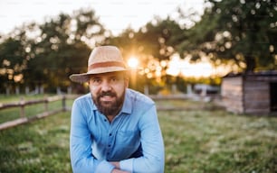 日没時に家族の農場で屋外に立つ帽子とエプロンを持つ幸せな成熟した男性の農家のポートレート。