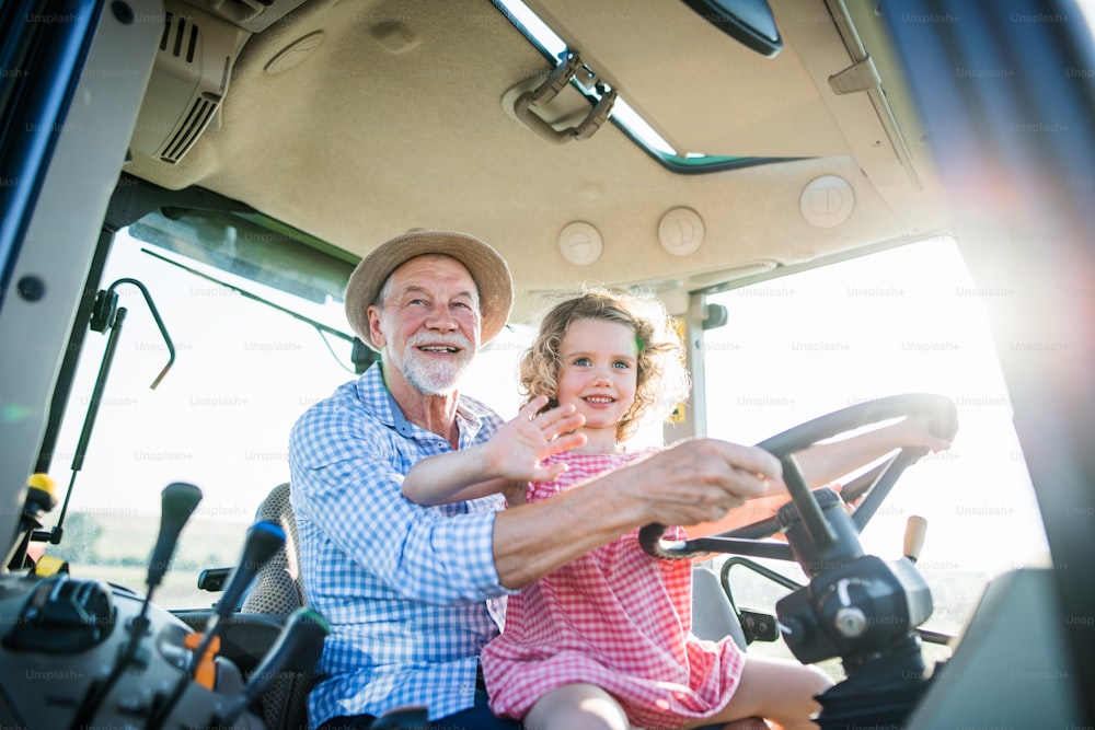 A senior farmer with small granddaughter sitting in tractor, driving.