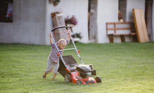 Down syndrome child with lawn mower walking outdoors in garden in summer.