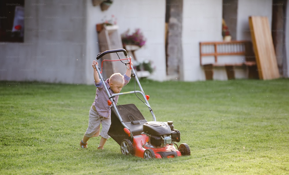 Niño con síndrome de Down con cortadora de césped caminando al aire libre en el jardín en verano.
