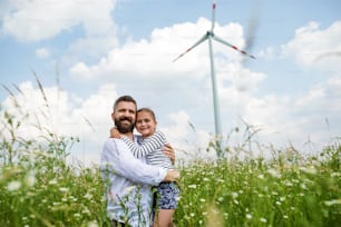 Padre maduro con hija pequeña de pie en el campo en un parque eólico, abrazándose.
