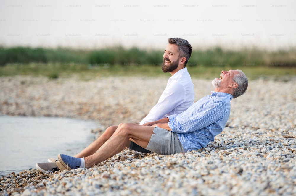 Padre mayor e hijo maduro sentados junto al lago, riendo. Espacio de copia.