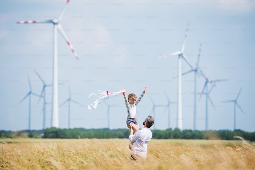 Mature father with small daughter standing on field on wind farm, playing.