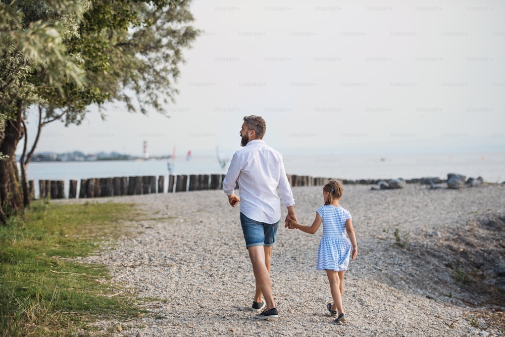 Vue arrière d’un père mûr marchant avec sa petite fille en vacances au bord du lac, se tenant la main.
