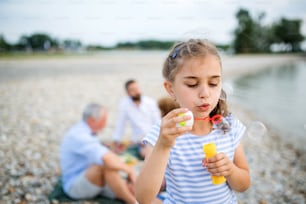 Front view of small girl with family on a holiday by the lake, making soap bubbles.