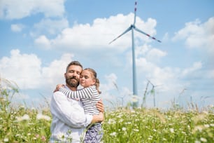 Mature father with small daughter standing on field on wind farm, eyes closed.
