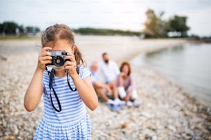 Vista frontal de una niña pequeña con cámara en unas vacaciones con la familia junto al lago, tomando fotos.