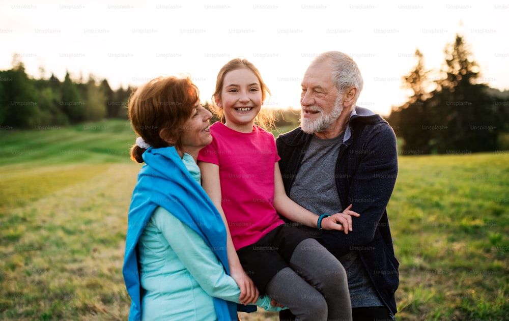 An active senior couple with cheerful granddaughter standing in nature at sunset.