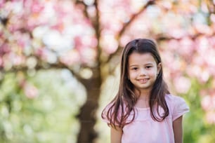 A cheerful small girl standing outside in spring nature, looking at camera.