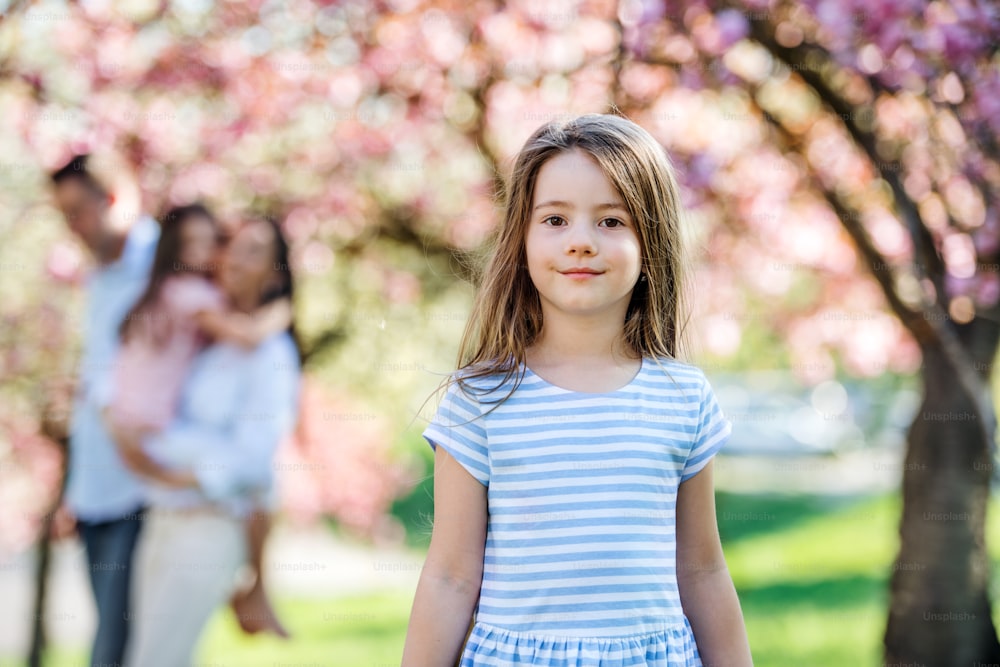 Eine Vorderansicht eines kleinen Mädchens mit Familie draußen in der Frühlingsnatur, mit Blick in die Kamera.