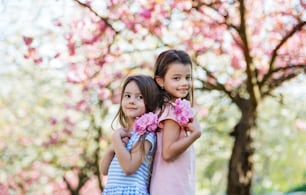 Zwei kleine Mädchen mit Blumen, die draußen in der Frühlingsnatur stehen und in die Kamera schauen.