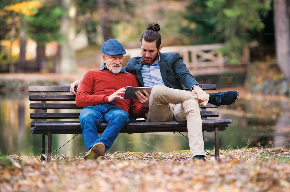Ein älterer Vater und sein Sohn sitzen auf einer Bank in der Natur und benutzen ein Tablet.