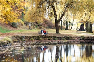 Pai sênior e seu filho pequeno sentados no banco à beira do lago na natureza, conversando.