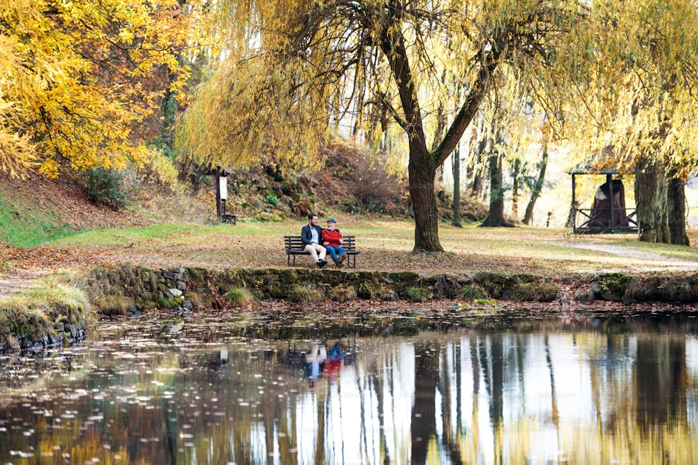 Un père aîné et son jeune fils assis sur un banc au bord du lac dans la nature, discutant.