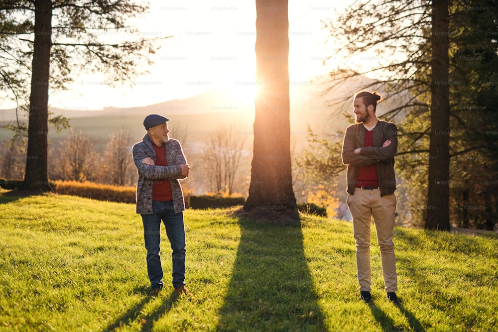 Senior father and his son with crossed arms standing by tree at sunset, looking at each other.