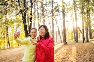Two female runners with smartphone standing outdoors in forest in autumn nature, taking selfie when resting.