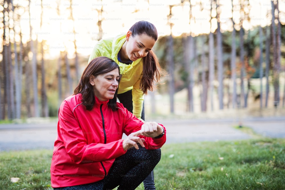 Two female runners with smartwatch standing on a road outdoors in forest in autumn nature, measuring or checking the time.