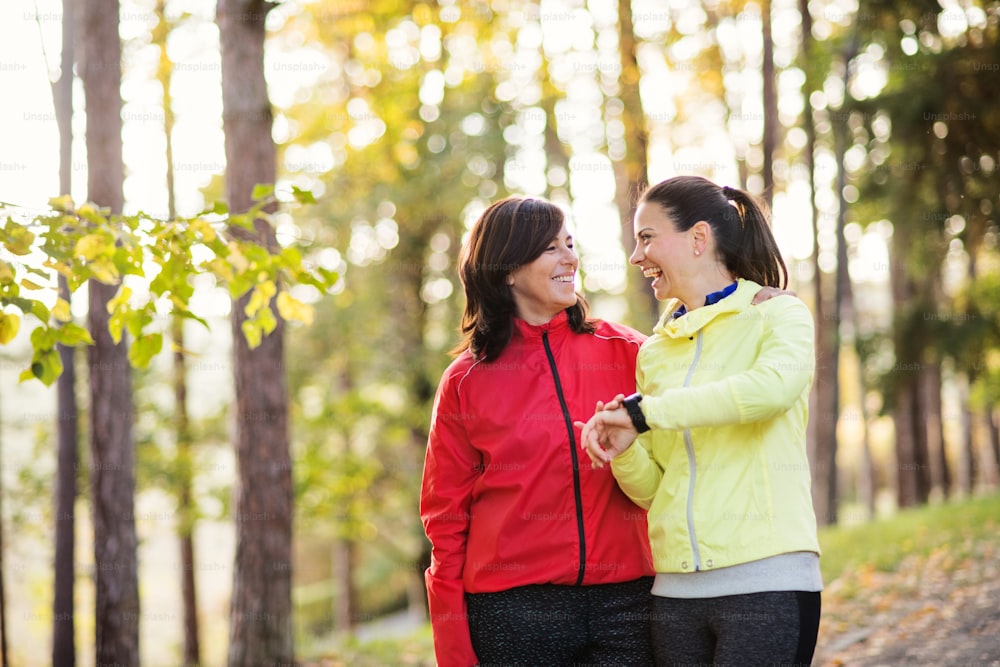 스마트워치를 든 두 명의 여성 주자가 가을 자연의 숲 속 야외 도로에 서서 시간을 측정하거나 확인한다.