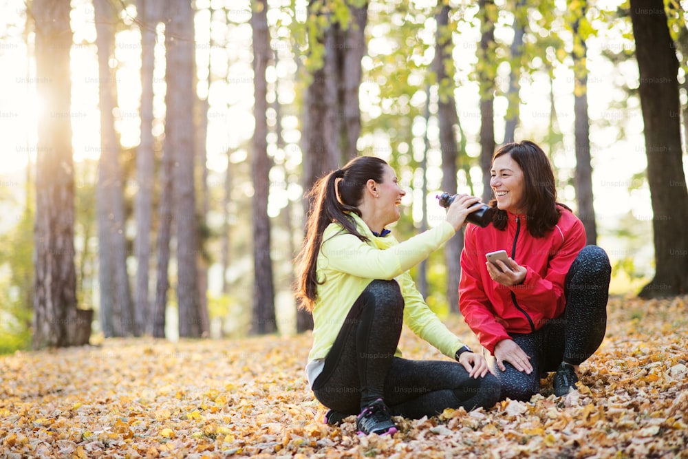 Two female runners with smartphone and water bottle sitting on the ground outdoors in forest in autumn nature, resting.