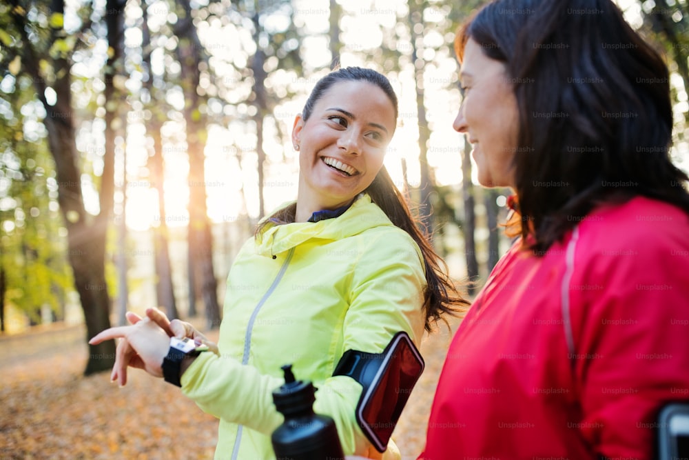 Two female runners with smartwatch and smartphone in arm band standing outdoors in forest in autumn nature, measuring or checking the time.