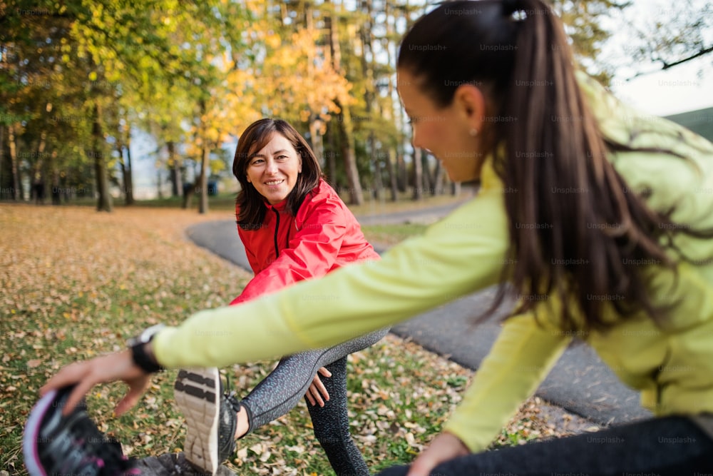 Two active female runners stretching legs outdoors in park in autumn nature after the run.