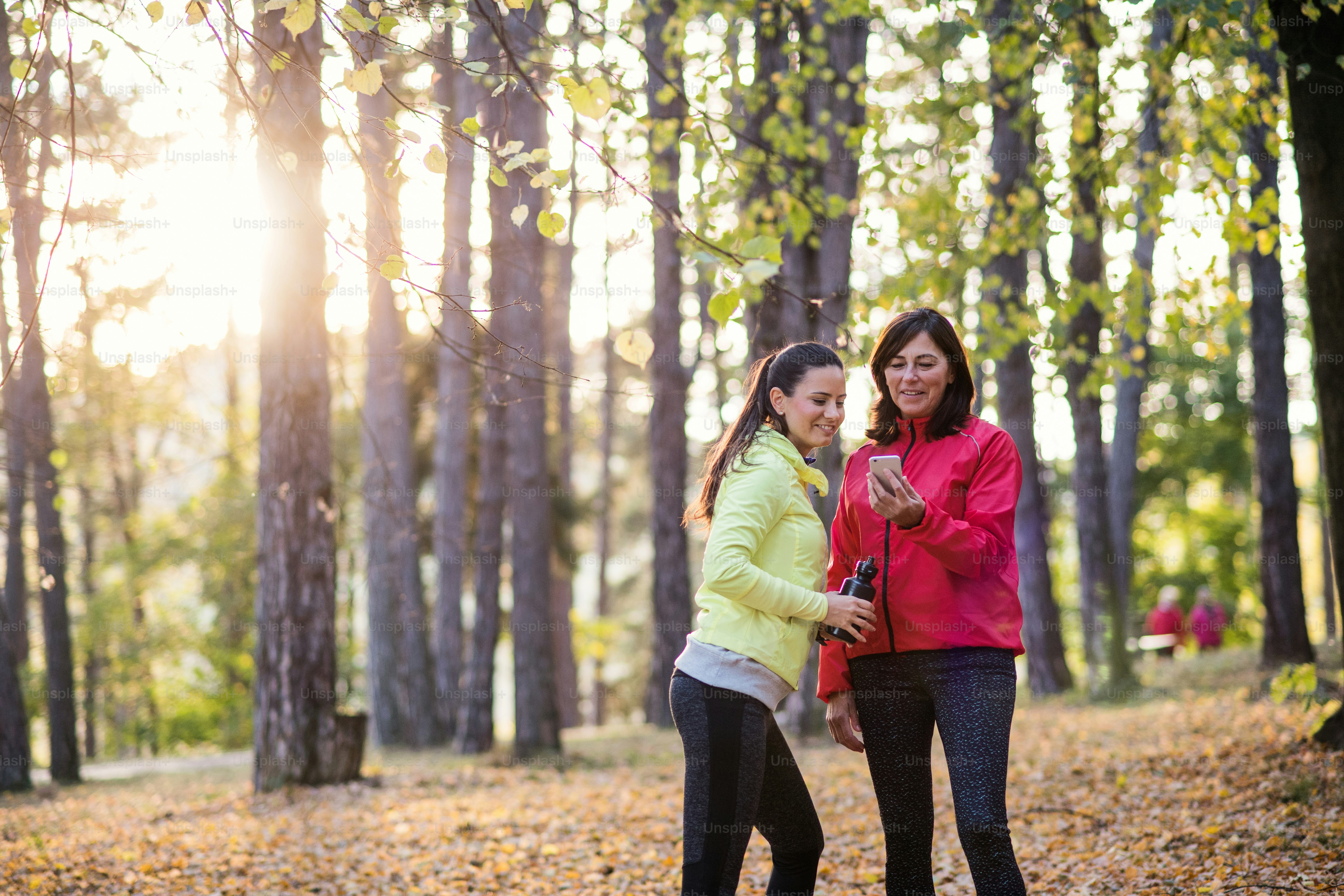 Zwei Läuferinnen stehen draußen im Wald in der Herbstnatur, mit Smartphone.