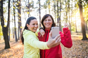 Two female runners with smartphone standing outdoors in forest in autumn nature, taking selfie when resting.