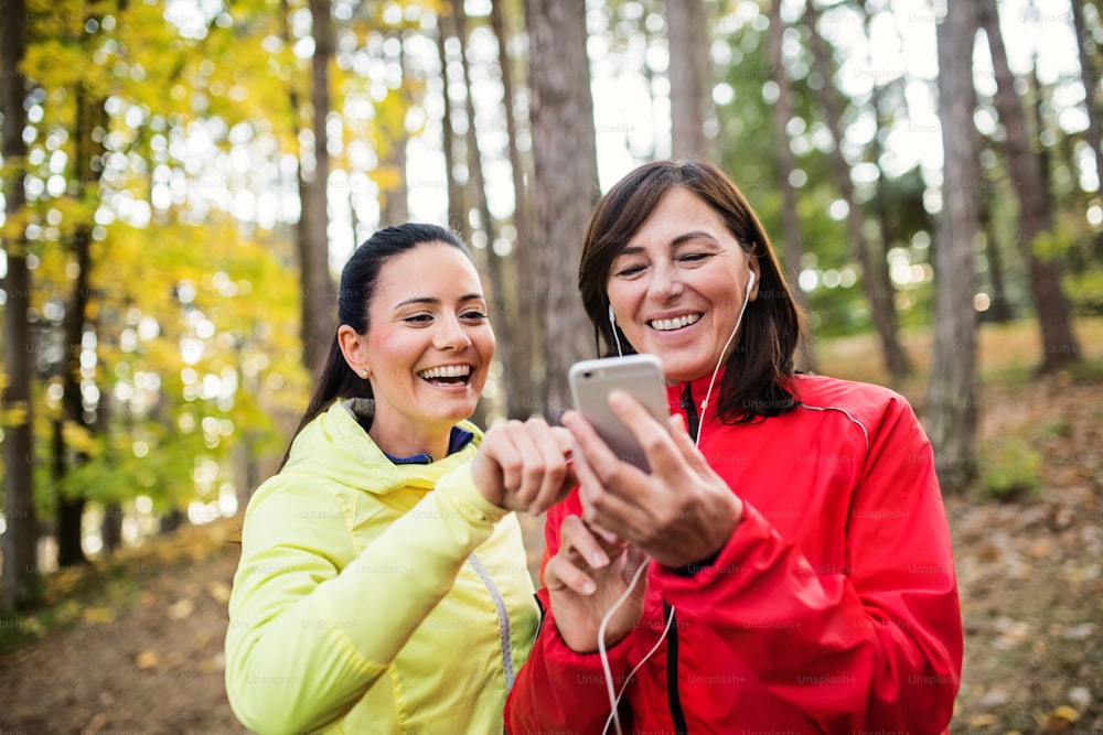Two female runners standing outdoors in forest in autumn nature, using smartphone.