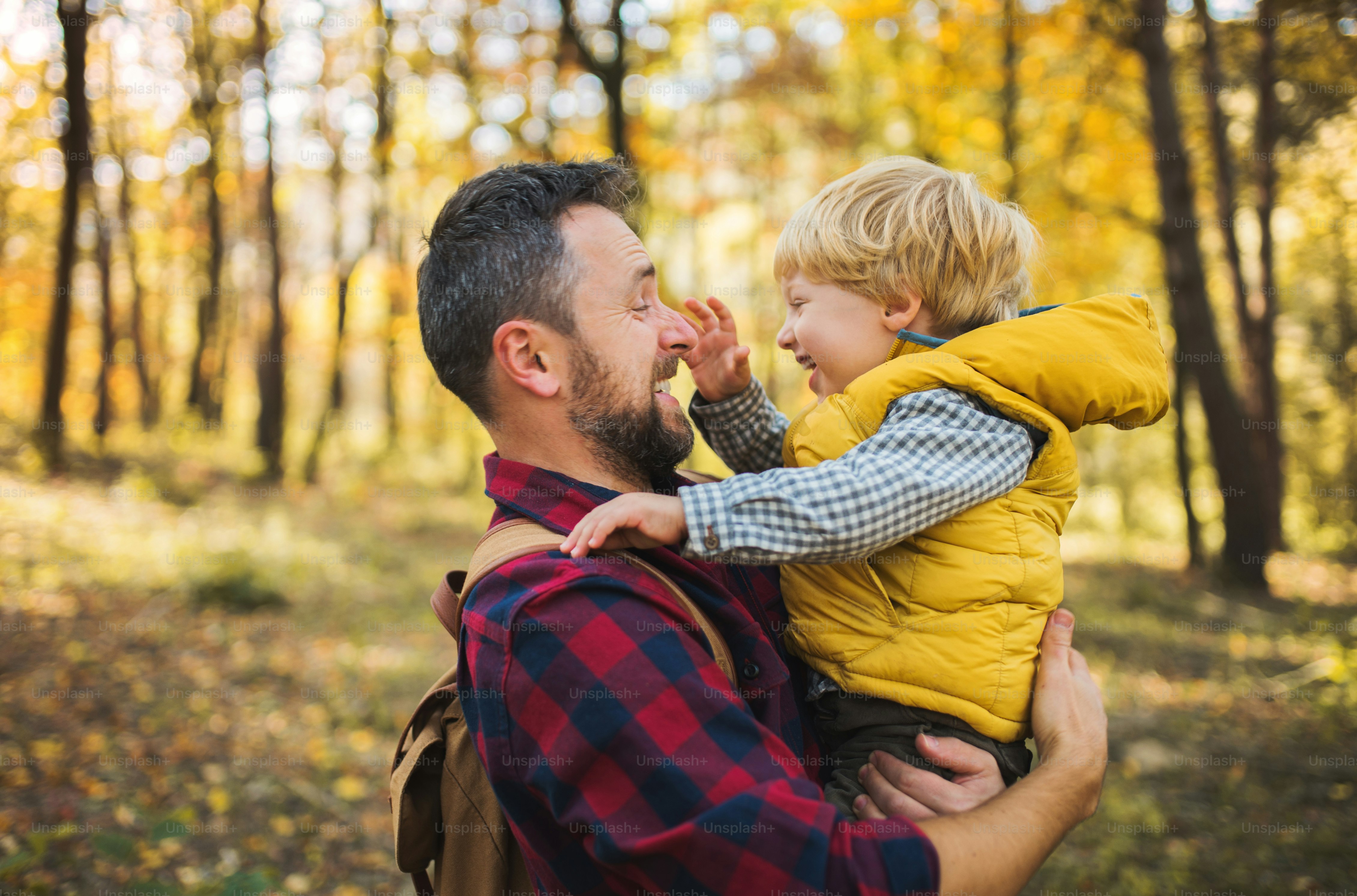 A mature father standing and holding a toddler son in an autumn forest, having fun.