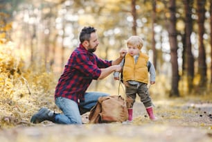 Um pai maduro colocando uma mochila em um filho pequeno em uma estrada em uma floresta de outono.