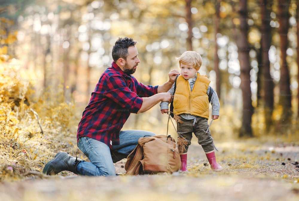 A mature father putting a backpack on a toddler son on a road in an autumn forest.