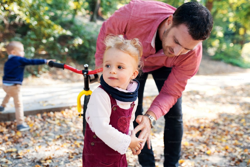 秋の森を散歩するバランスバイクを持つ父親と小さな双子のポートレート。