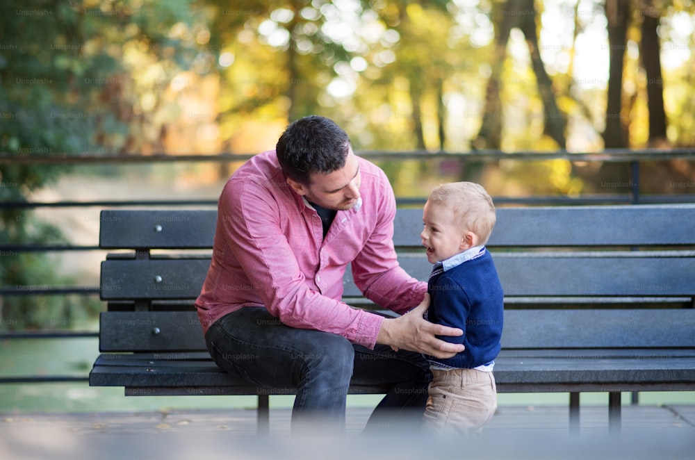 A father with small son sitting on bench in park in autumn.