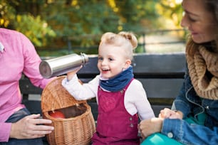 A beautiful young family and small daugther with picnic basket in autumn park, sitting on bench.