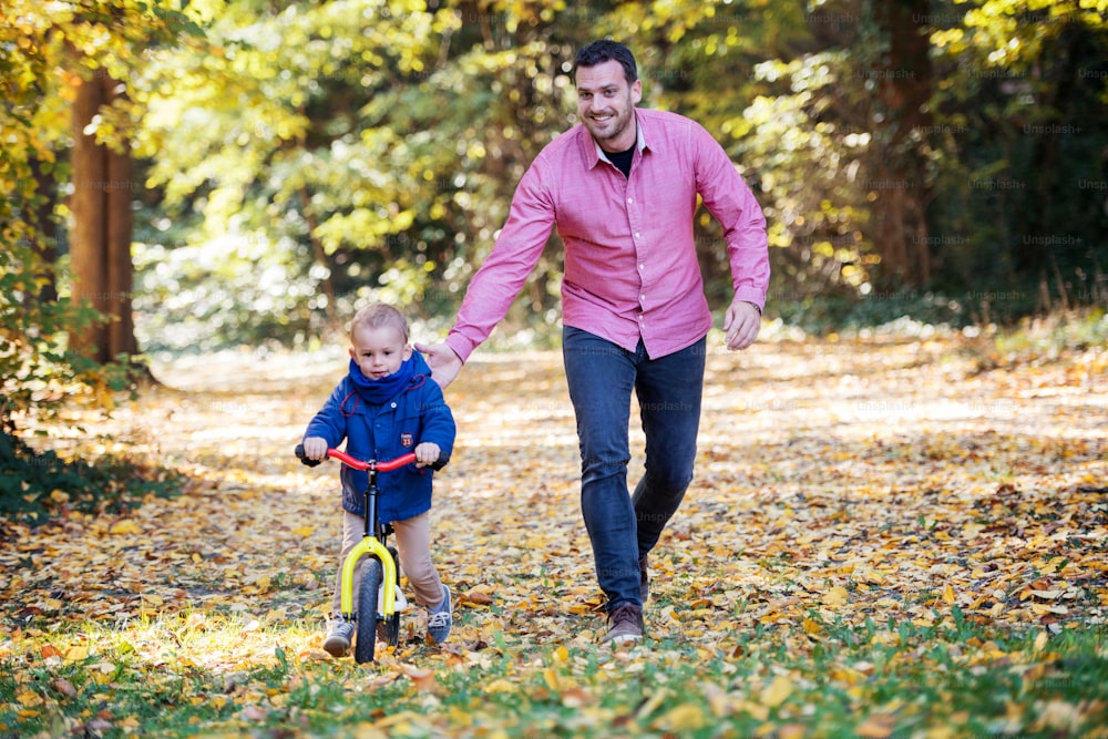 Front view of father and small son with a balance bike on a walk in autumn forest.