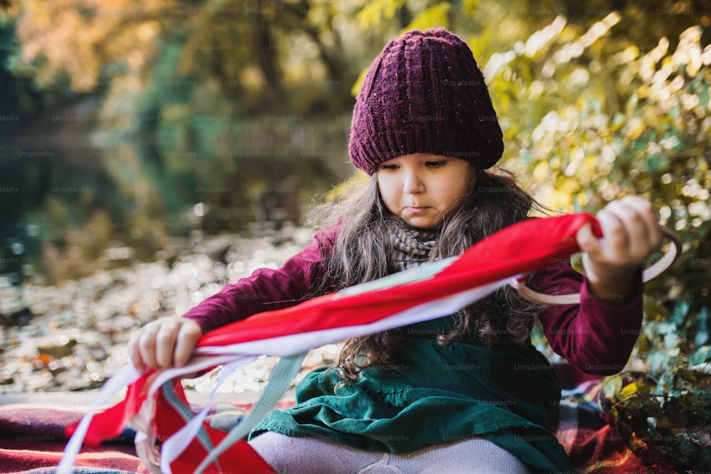 A portrait of a small toddler girl sitting in forest in autumn nature playing with ribbon kite.