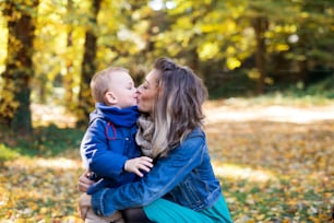 Beautiful young mother with small toddler son on a walk in autumn forest, kissing.