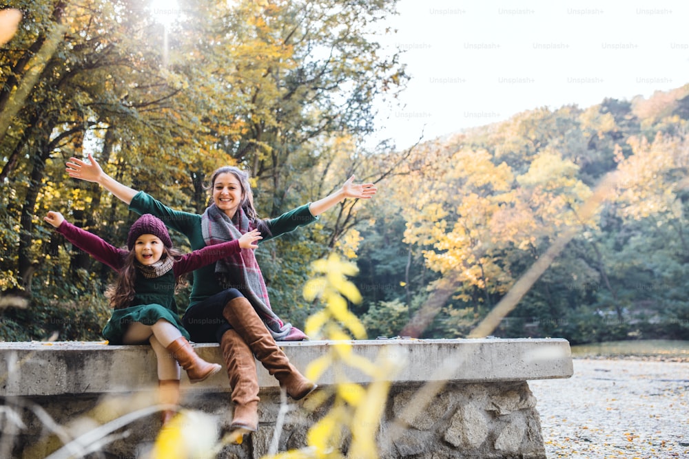 A mother with a toddler daughter sitting on wall in forest in autumn nature, stretching arms.