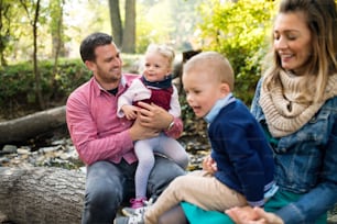 A beautiful young family with small twins on a walk in autumn forest, sitting on log.