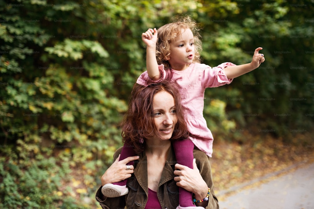 Beautiful young mother with toddler daughter on a walk in autumn forest, giving her piggyback ride.