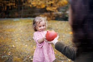 Small toddler girl with unrecognizable mother on a walk in autumn forest, holding hokkaido pumpkin.