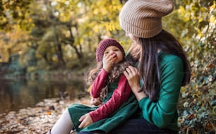 A portrait of young mother with a toddler daughter hugging and kissing in forest in autumn nature.