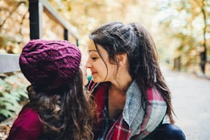 A portrait of young mother with a toddler daughter touching noses in forest in autumn nature.