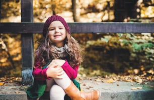 A portrait of a small toddler girl sitting with crossed legs in forest in autumn nature.