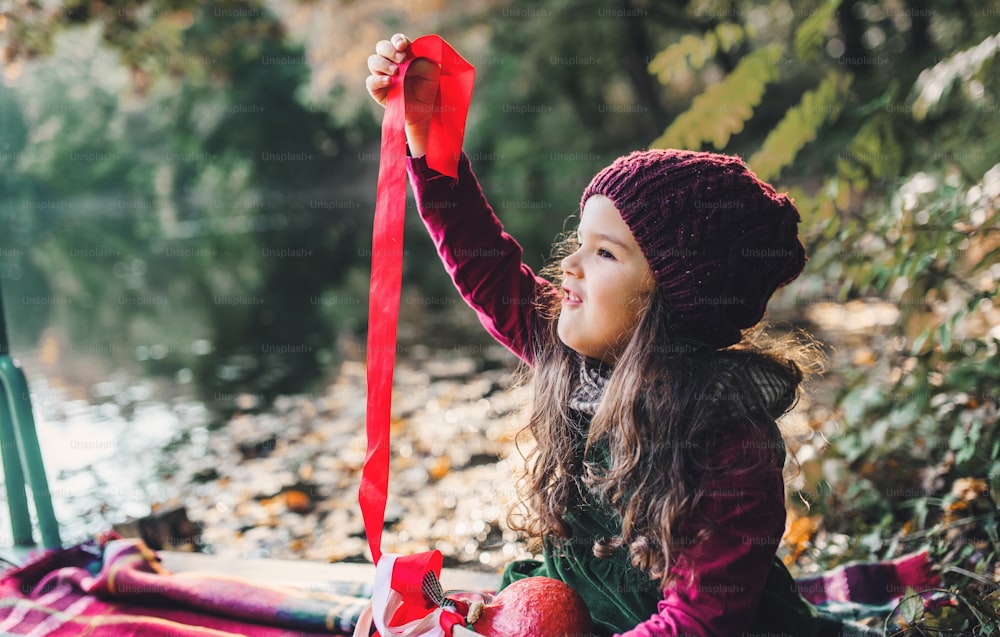 A portrait of a small toddler girl sitting in forest in autumn nature playing with ribbons.
