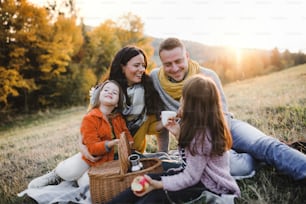 Ein Porträt einer glücklichen jungen Familie mit zwei kleinen Kindern, die bei Sonnenuntergang in der Herbstnatur auf einem Boden sitzen und ein Picknick machen.