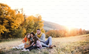 A portrait of happy young family with two small children sitting on a ground in autumn nature at sunset, having picnic.
