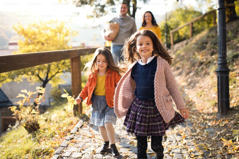Duas meninas com pais irreconhecíveis ao fundo andando no parque no outono, de mãos dadas.